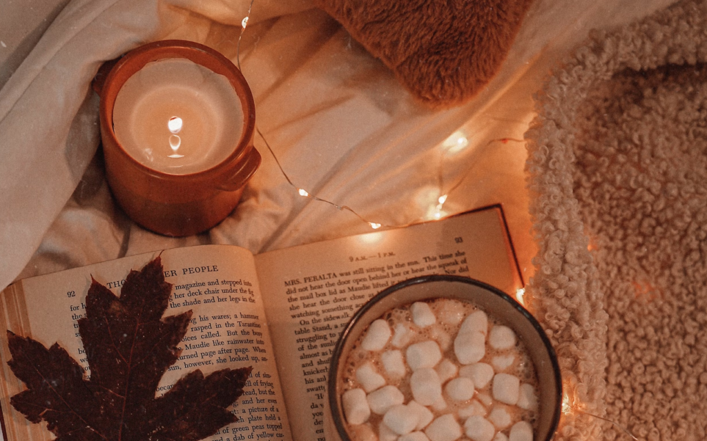 A photograph of a cozy autumn still life featuring a fallen leaf, cocoa with marshmallows, an open book, a candle, and string lights white atop bedsheets.